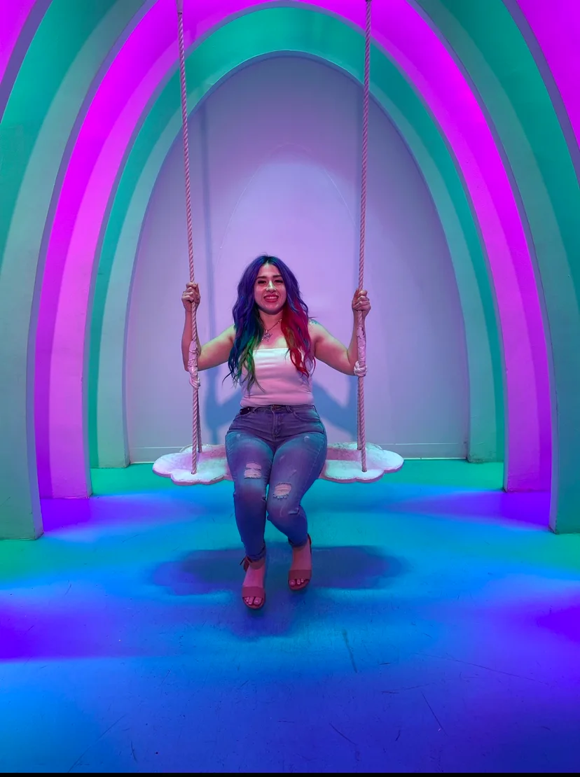 A Sueños Salon stylist with vivid multicolor hair seated in a glowing teal and magenta arched room